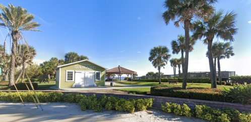 A sunny beach scene featuring a green garage, gazebo, palm trees, and well-maintained landscaping.