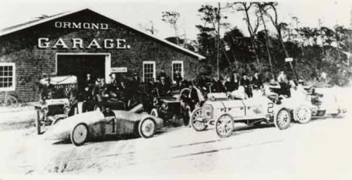 Historic photo of early race cars and drivers outside the Ormond Garage, surrounded by trees.