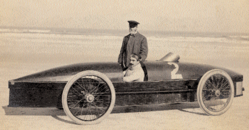 Two men pose beside a vintage racing car on a beach, with one seated inside and the other standing on top.