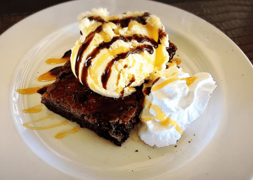 A decadent brownie topped with vanilla ice cream, chocolate drizzle, whipped cream, and caramel sauce on a white plate.