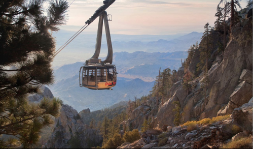 A cable car glides over rocky terrain, surrounded by trees and mountains, with a vast landscape in the background.