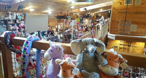 A cozy shop interior filled with stuffed animals and toys, viewed from a balcony with colorful plush toys in the foreground.