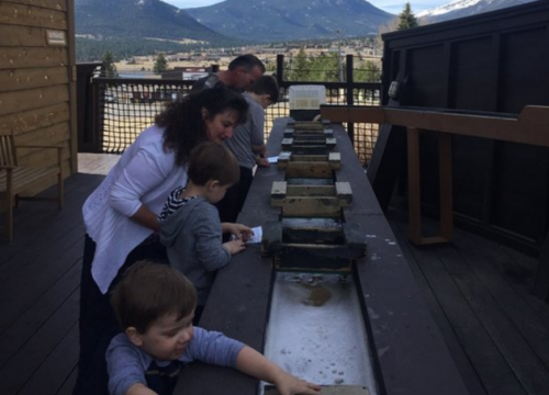 A family with two young children explores a water feature outdoors, surrounded by mountains and wooden structures.
