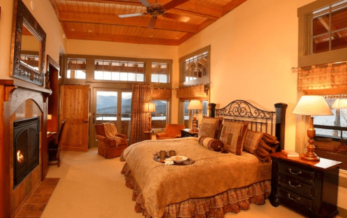 Cozy bedroom with a fireplace, large windows, and warm decor, featuring a bed with a patterned blanket and a seating area.