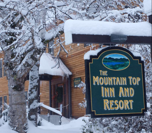 A snow-covered sign for the Mountain Top Inn and Resort, with a wooden building and trees in the background.