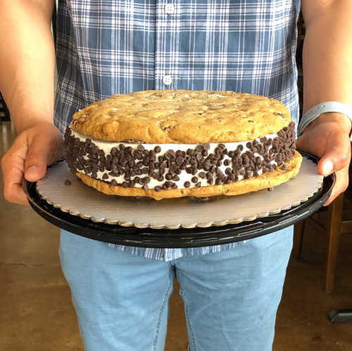 A person holds a large cookie sandwich filled with ice cream and chocolate chips on a black serving tray.