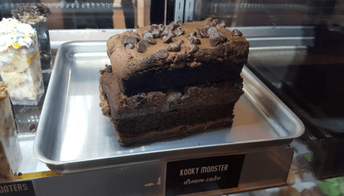 A layered chocolate cake topped with chocolate chips, displayed in a bakery case with a label "Kooky Monster dream cake."