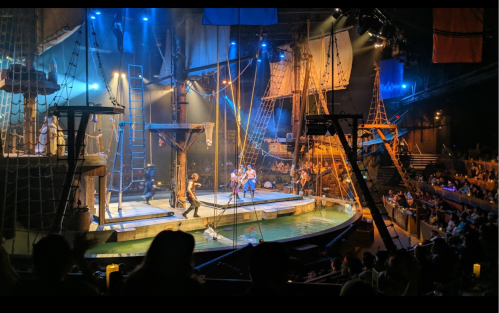A lively stage performance on a ship-themed set, with actors and a water-filled arena, surrounded by an audience.