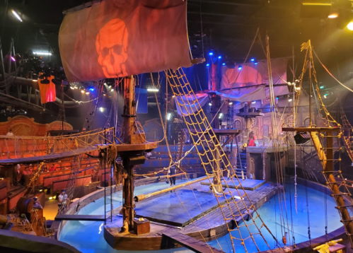 A pirate-themed stage with a ship, skull flag, and water, illuminated by colorful lights in a lively atmosphere.
