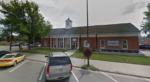 A brick building with a clock tower, featuring a health center sign, surrounded by trees and parked cars.