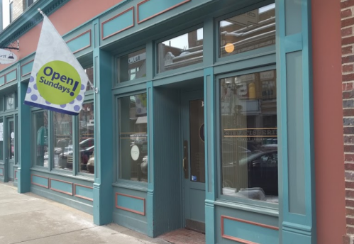 Storefront with teal trim and a flag reading "Open Sundays" hanging outside. Cars parked nearby.