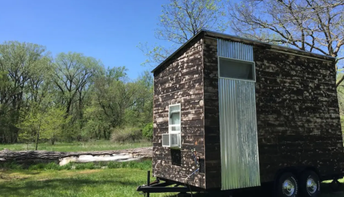 A rustic tiny house on wheels, set in a green landscape with trees and a clear blue sky.