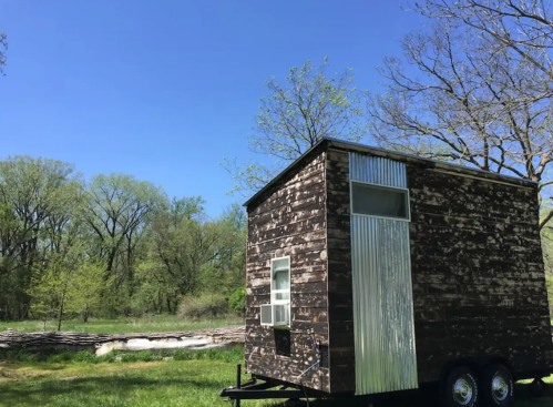 A small wooden tiny house on wheels, set in a green landscape under a clear blue sky.