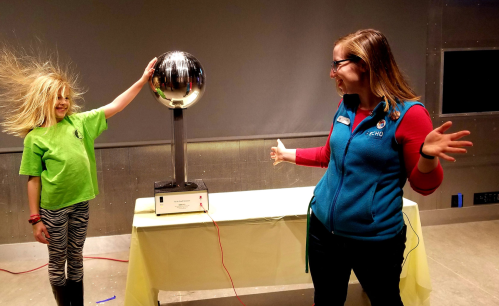 A child with wild hair stands next to a Tesla coil, while an adult in a blue vest gestures excitedly.