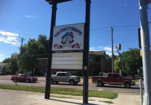 Sign for "The Donut Shoppe" featuring a cartoon donut character, with cars passing by on a sunny day.
