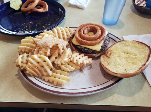 A plate with a cheeseburger topped with onion rings, served with waffle fries and a toasted bun on the side.