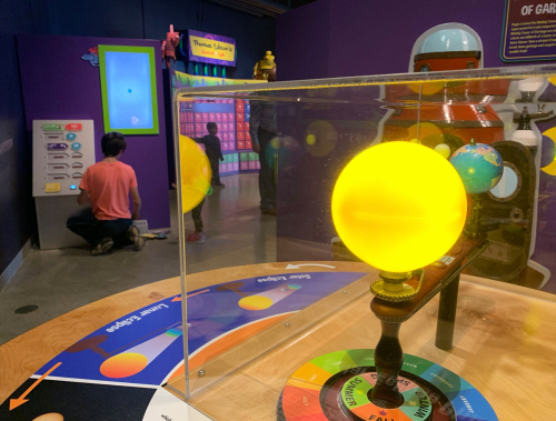A glowing yellow model of the sun in a science exhibit, with children interacting with displays in the background.