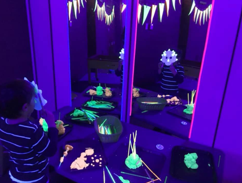 A child holds a mask in a dark room with glowing playdough and decorations, reflected in multiple mirrors.