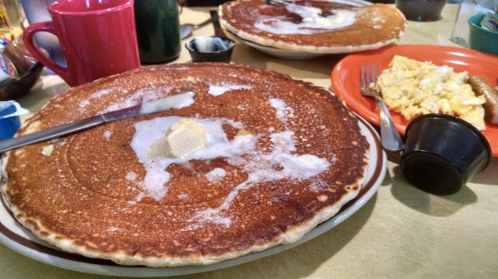 A large pancake with melted butter on a plate, alongside a plate of scrambled eggs and a red mug.