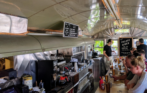 Interior of a diner with a menu board, a barista serving coffee, and customers seated at the counter.