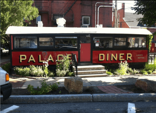 A vintage diner with a red and black exterior, featuring the sign "Palace Diner" and surrounded by greenery.