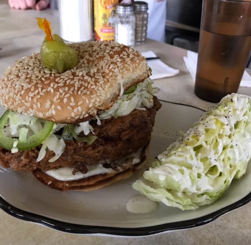 A large sesame seed bun sandwich filled with fried chicken, lettuce, and jalapeños, served with a side of lettuce.