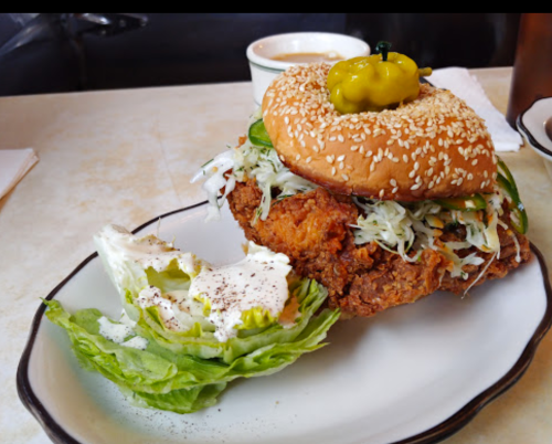 A sesame seed bun sandwich with fried chicken, topped with slaw and jalapeños, served with a side of lettuce.