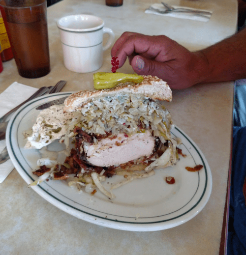 A large sandwich with fried chicken, coleslaw, and a pickle on top, served on a plate with utensils nearby.