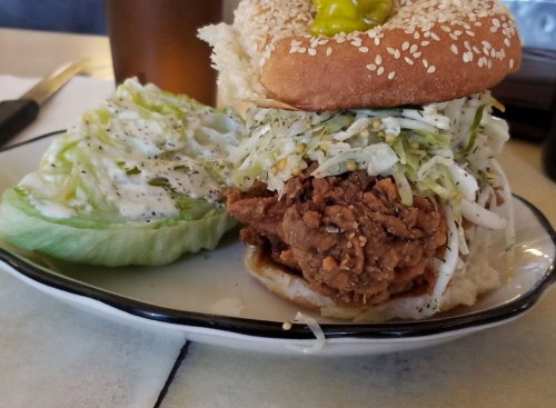 A sesame seed bun sandwich with fried chicken, coleslaw, and a pickle, served with a side of lettuce.