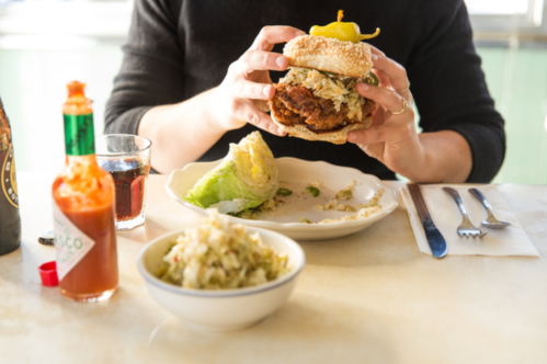 A person holds a large sandwich filled with coleslaw, with a side of coleslaw and a drink on the table.