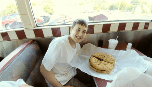 A smiling boy sits at a table with a large sandwich wrapped in paper in front of him.