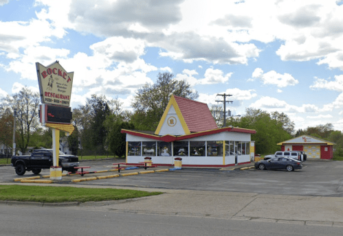 A colorful fast-food restaurant with a red and yellow roof, surrounded by trees and parked cars under a cloudy sky.