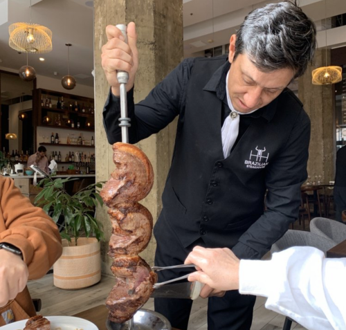 A waiter carves skewered meat at a restaurant, serving it to a diner at the table.