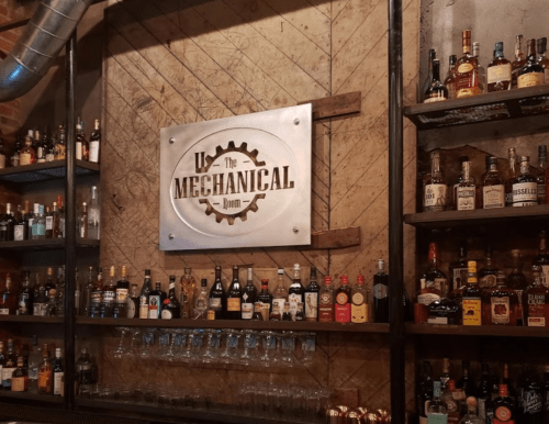 A bar with a sign reading "The Mechanical Room" and shelves stocked with various bottles of liquor.