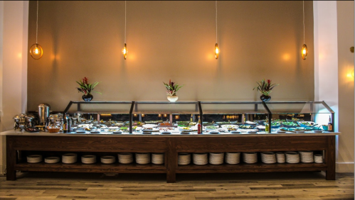 A buffet table with a glass display showcasing various dishes, surrounded by decorative plants and pendant lights.