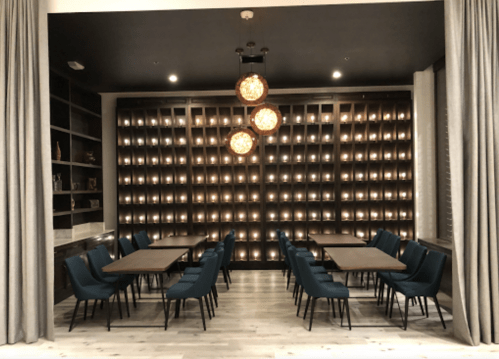 A modern dining area with blue chairs, wooden tables, and a wall of lit candles, framed by elegant curtains.
