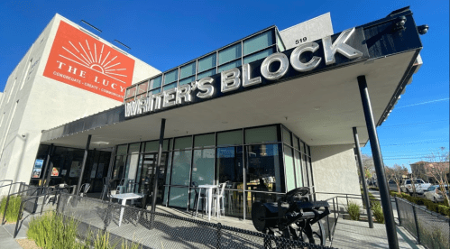 Exterior of a modern building featuring "Writer's Block" sign, with outdoor seating and a bright blue sky above.