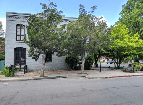A gray building with trees in front, located on a quiet street with a few parked cars and greenery nearby.