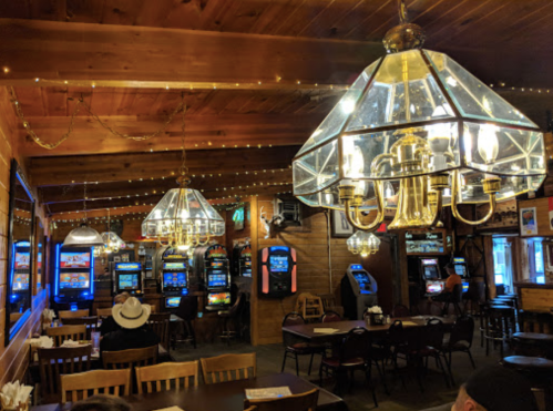 Cozy interior of a rustic restaurant with slot machines, wooden tables, and warm lighting from chandeliers.
