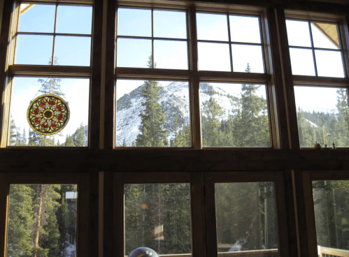 View from a cabin window showcasing a mountain and tall pine trees, with a decorative stained glass hanging.