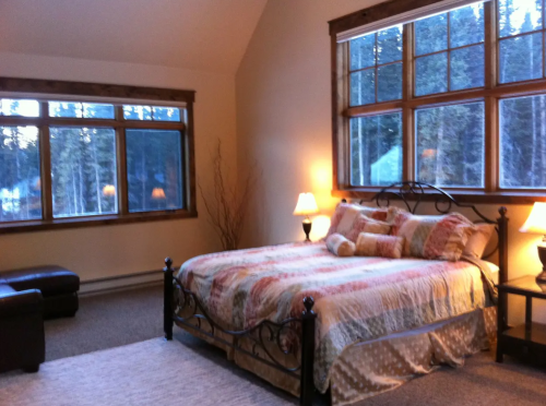 Cozy bedroom with a large bed, warm lighting, and windows overlooking a forested area.