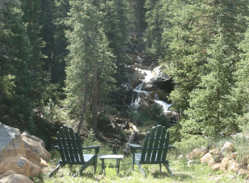 Two green chairs and a small table sit beside a flowing stream, surrounded by lush trees and rocky terrain.