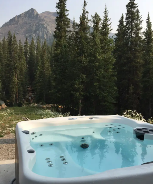 A hot tub sits on a patio surrounded by tall pine trees and mountains in the background, under a clear sky.