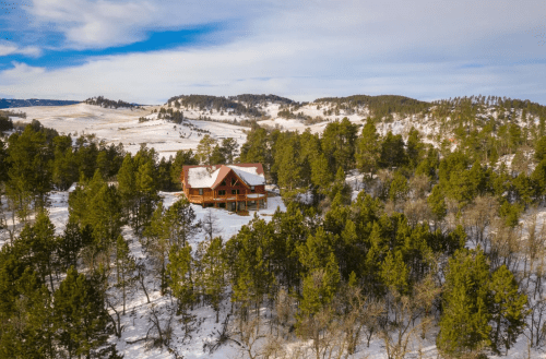 A large wooden cabin surrounded by snow-covered trees and rolling hills under a clear blue sky.