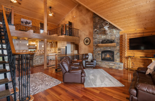 Cozy cabin interior with wooden beams, stone fireplace, leather chairs, and open kitchen area. Warm and inviting atmosphere.