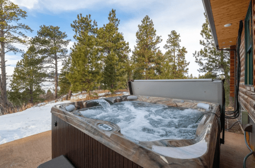 A hot tub with bubbling water, surrounded by snow and tall pine trees under a blue sky.
