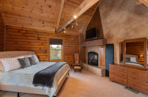 Cozy bedroom with wooden walls, a large bed, fireplace, TV, and rustic decor, featuring a mountain view.