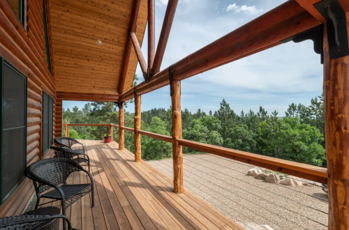 A wooden deck with black chairs overlooks a lush green landscape and trees under a partly cloudy sky.