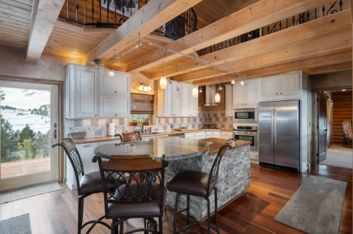 Modern kitchen with stone accents, wooden beams, and a large island with bar seating, overlooking a scenic view.