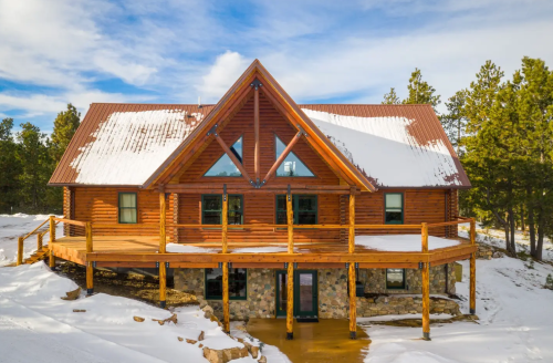 A large log cabin with a triangular roof, surrounded by snow and trees, featuring a spacious wooden deck.